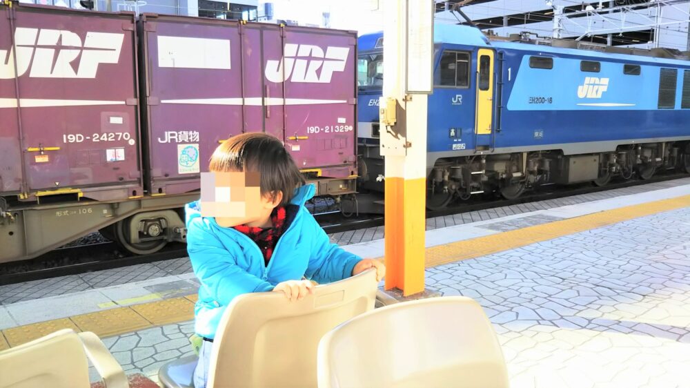 ＪＲ王子駅の中央線松本方面側ホームで見える貨物列車と、ベンチに座って電車を見る子供