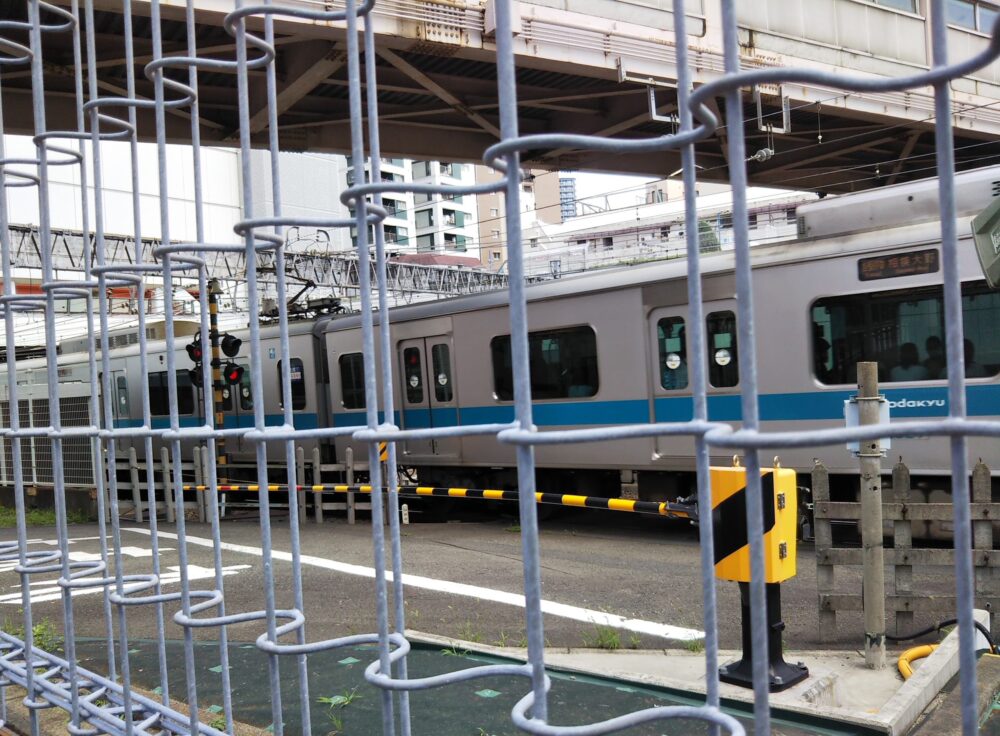 相模大野駅にある車両基地前の線路沿い広場で見える、フェンス内にある踏切と踏切を通過する電車